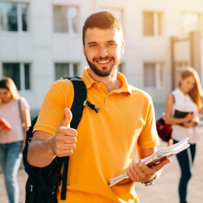 Young attractive smiling student showing thumb up outdoors on campus at the university. Selective focus